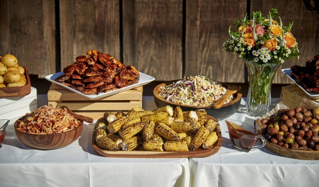 A well-arranged catering table with a selection of dishes including roasted potatoes, chargrilled corn, a large portion of chicken wings, pasta, coleslaw, seasoned bread rolls, and a vase of fresh flowers, all set against a wooden fence for an outdoor event.