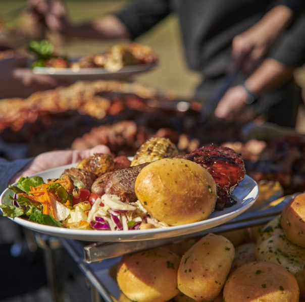A plate held by a person featuring a variety of barbecue foods including a fresh garden salad, grilled sausage, a roll, and barbecued chicken thigh, with more food being served in the background.