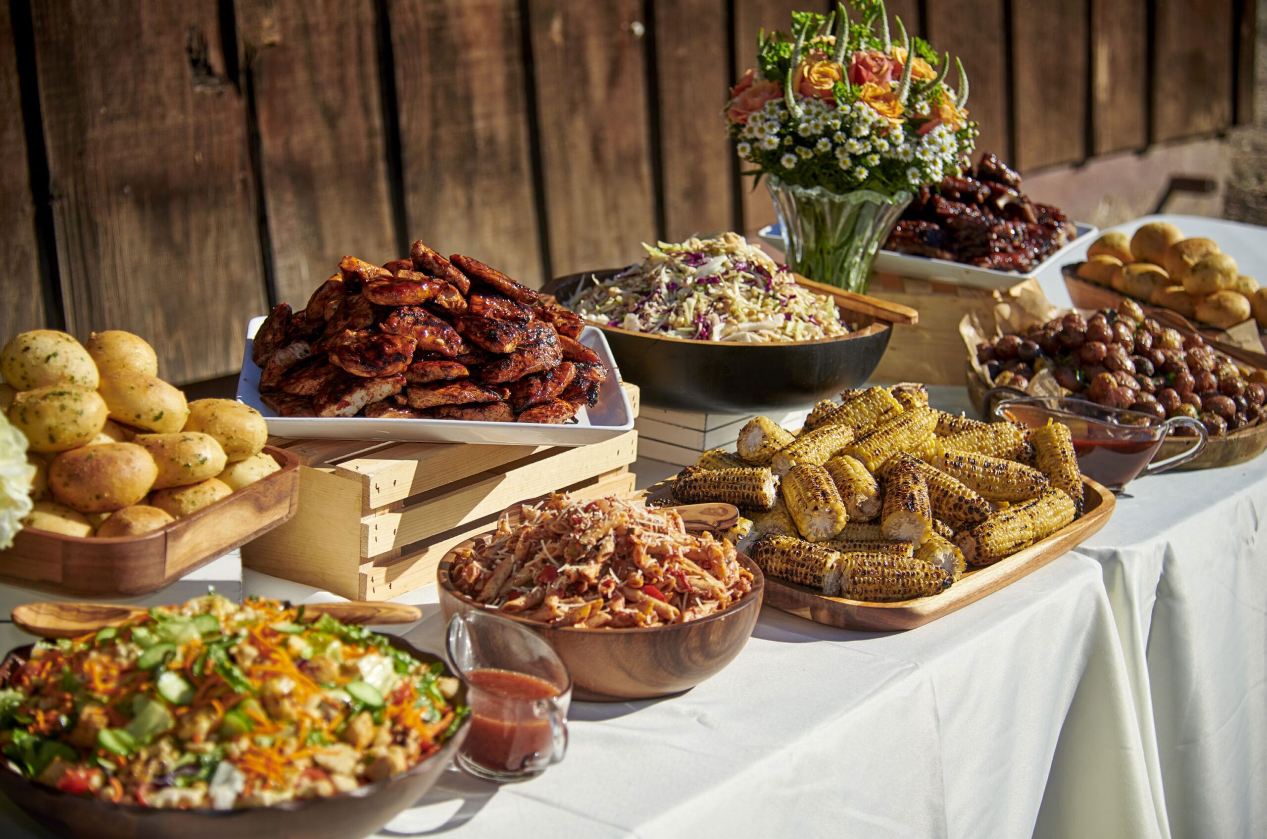 An outdoor catering event setup with a rustic wooden backdrop featuring dishes such as chargrilled chicken wings, pulled pork, corn on the cob, a colorful salad, baked rolls, coleslaw, and a bouquet of flowers adding a decorative touch to the bountiful spread.