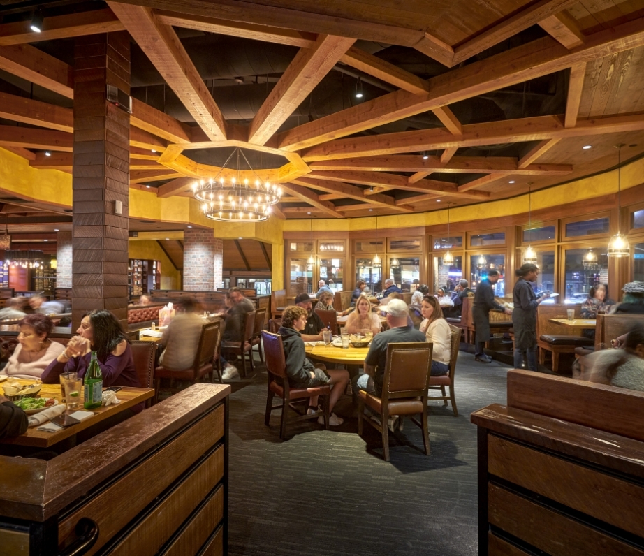 A spacious restaurant dining area filled with patrons, featuring an elegant chandelier, exposed wooden beams, and a warm, inviting atmosphere.