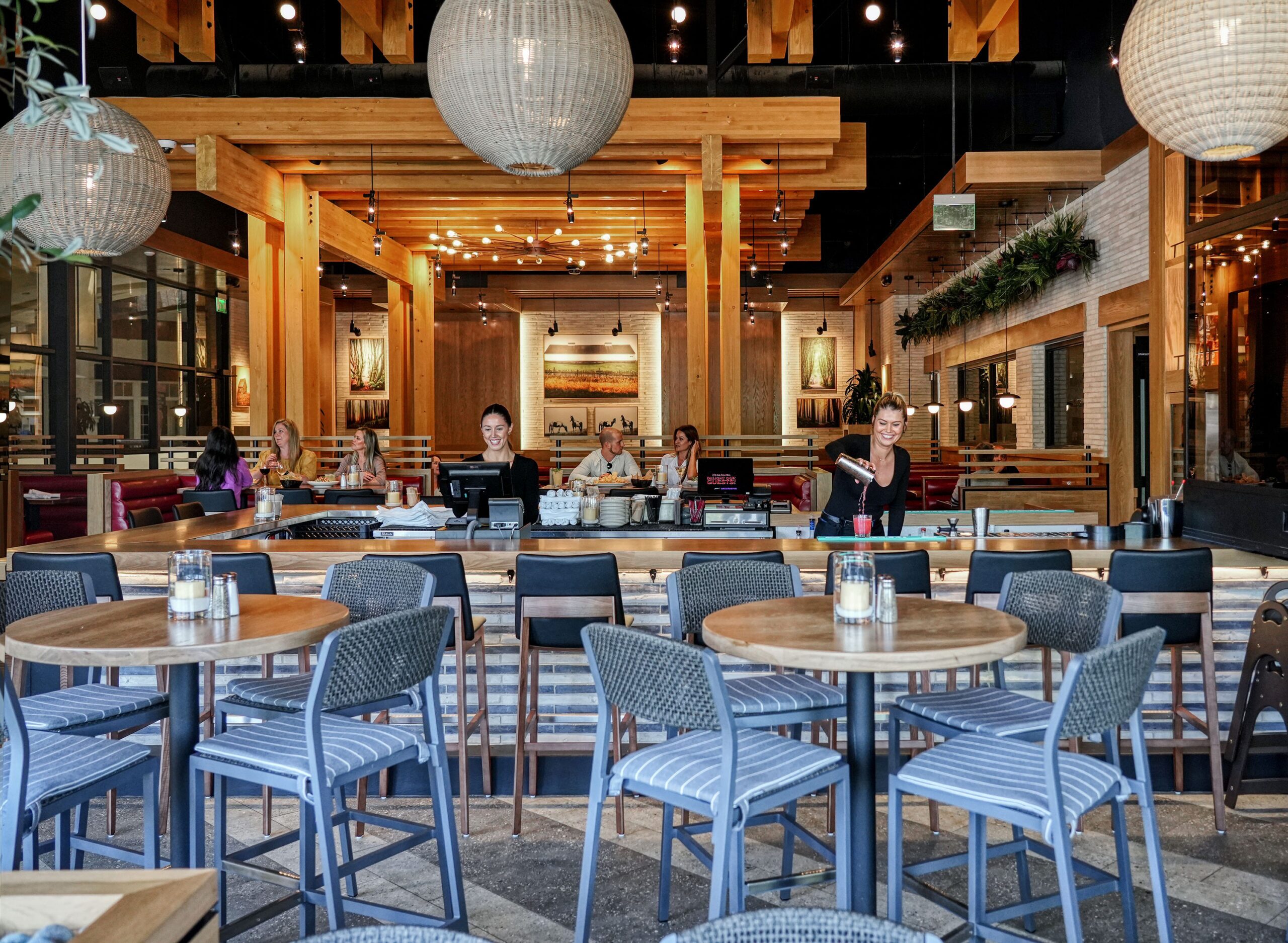 pacious and inviting restaurant interior with natural wood structures, large woven globe pendant lights, and a mix of booth and chair seating. Customers are dining in the background, and friendly staff are present, creating a welcoming atmosphere.
