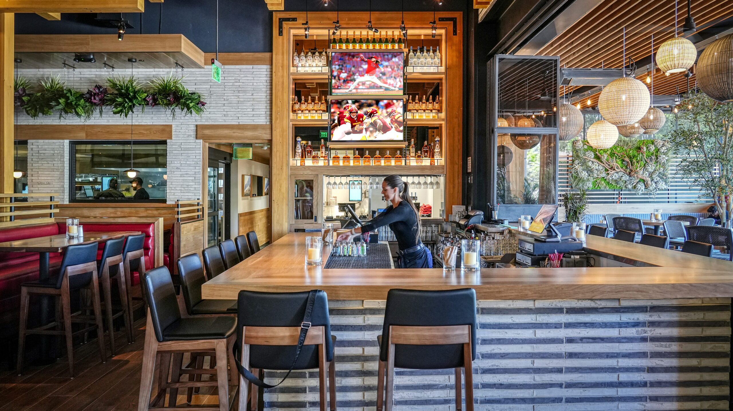 A modern restaurant bar area with a bartender at work, featuring a sports game on the television screens, wooden beams overhead, and hanging spherical lanterns, creating a contemporary and inviting ambiance.