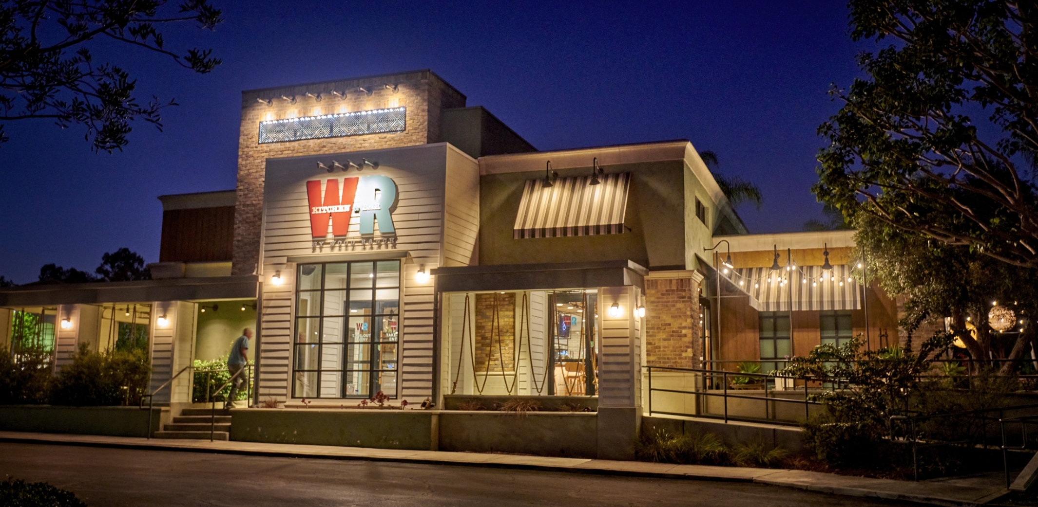 The inviting entrance of Wood Ranch Kitchen & Bar at twilight, with its welcoming lights, modern structure, and a well-manicured outdoor seating area.