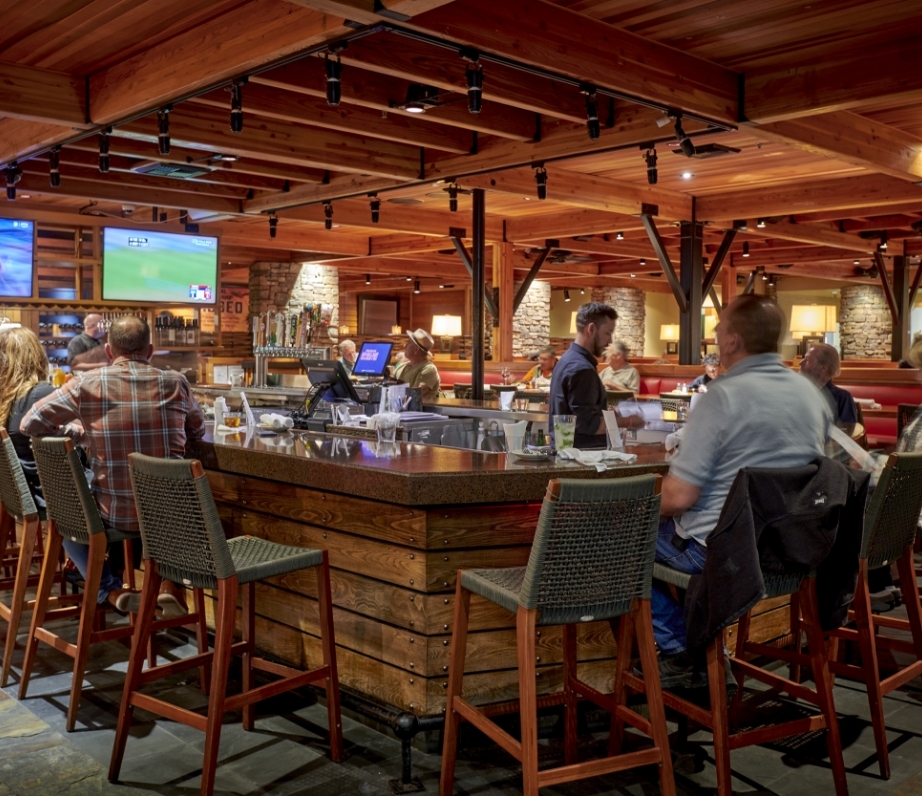 Patrons seated at the bar in Wood Ranch BBQ & Grill, Ventura, enjoying a convivial atmosphere with overhead sports telecasts and a rustic, wood-dominated décor.