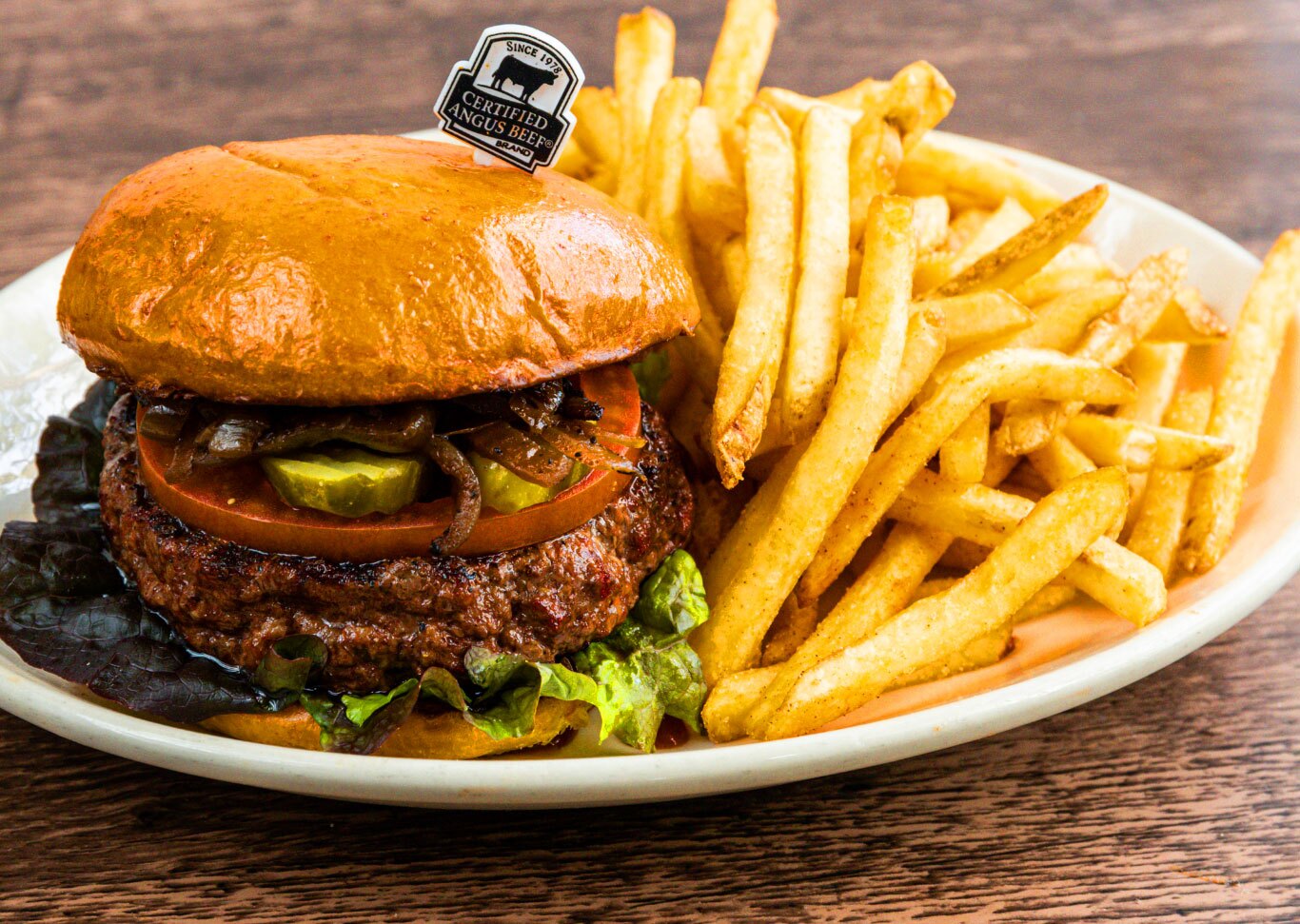 A plump hamburger with a shiny brioche bun, certified Angus beef patty, lettuce, tomato, pickles, and caramelized onions, accompanied by a side of golden French fries, served on a white plate with a wooden table background.