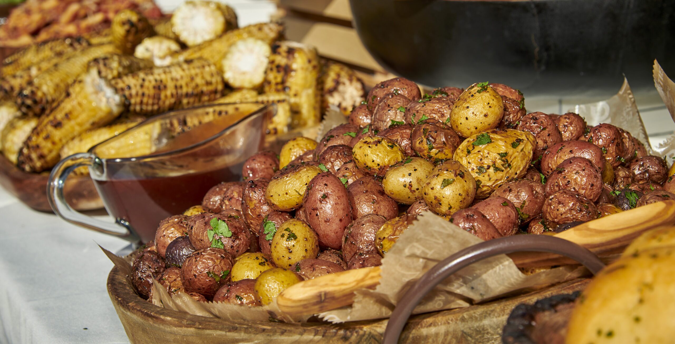 A wooden table holds a white ceramic bowl filled with small, round potatoes. Their skins are a mix of brown and yellow, and they appear to be roasted or baked. Next to the bowl sits a white ceramic pitcher with a blue rim. It is unclear what is inside the pitcher, but it could be gravy, sauce, or another condiment.