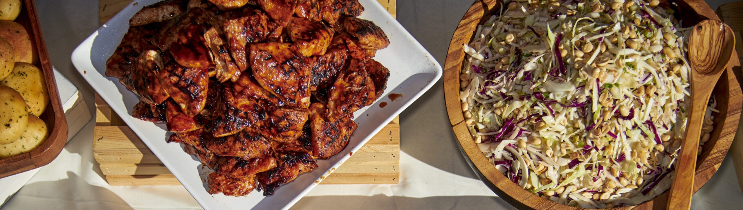 A panoramic view of a banquet table with a variety of dishes including seasoned rolls, a platter of grilled chicken, and a large bowl of Asian-inspired slaw sprinkled with peanuts, all under natural sunlight.