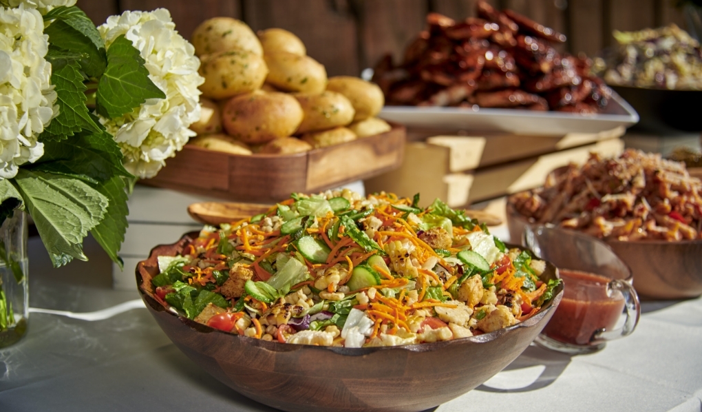 A buffet table set outdoors with a variety of dishes including a large colorful mixed salad in a wooden bowl, seasoned potatoes, barbecued chicken wings, and pasta, with a vase of white hydrangeas adding a touch of elegance to the display.