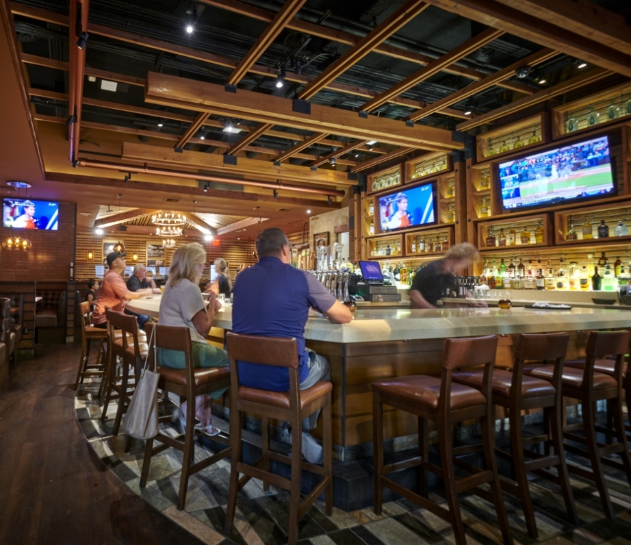 Guests at the full bar at Wood Ranch Irvine watching multiple sports broadcasts on overhead TVs, with warm wood finishes and amber lighting.