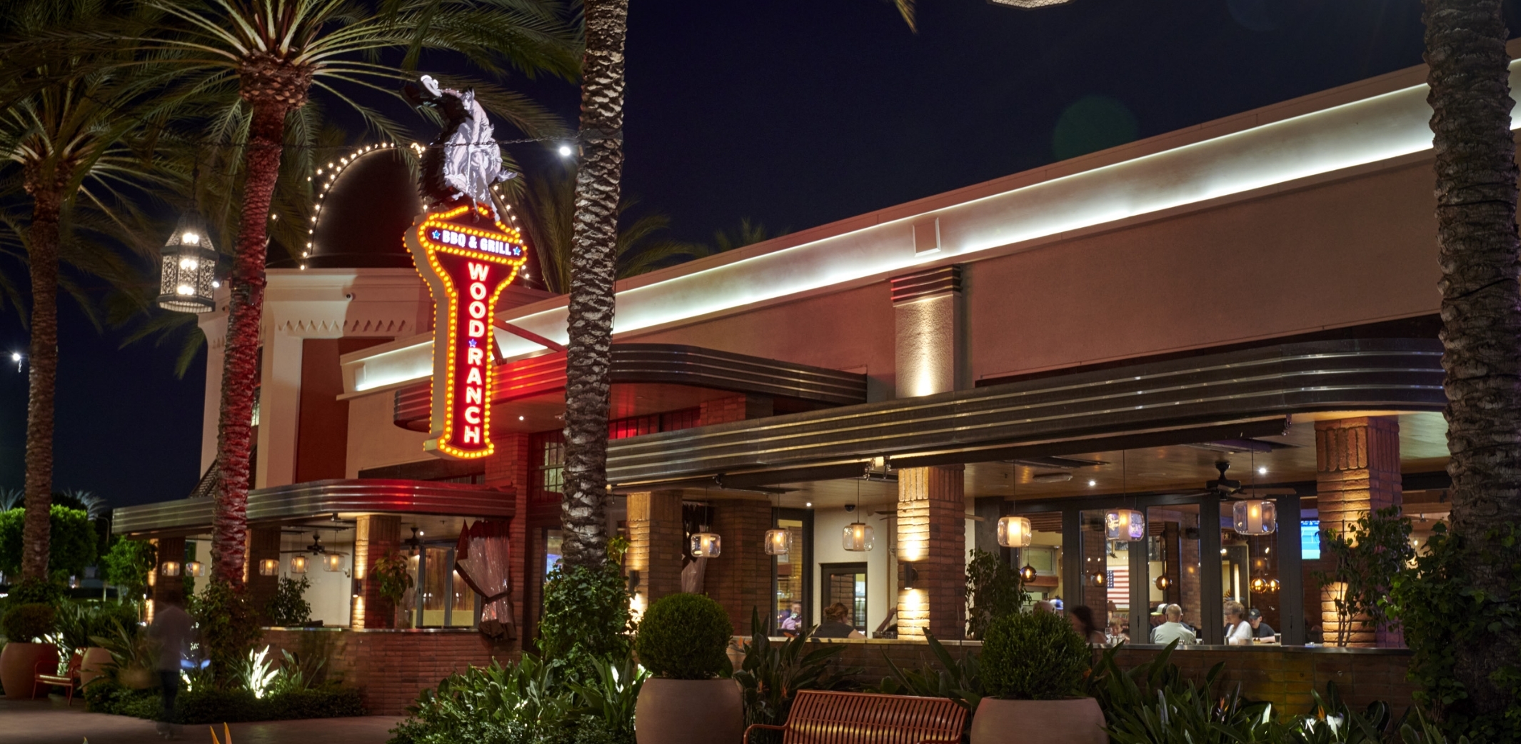 The Wood Ranch BBQ & Grill Irvine entrance at dusk, lit by warm neon signage and framed by tall palm trees at Spectrum Center.